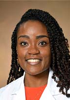 Headshot of woman with long dark braids, a doctor's white coat and an orange shirt. She is smiling.
