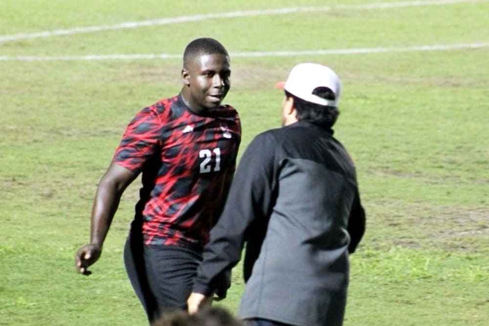 Young man in red and black soccer uniform with the number 21 on the shirt talks to a man wearing a baseball cap.
