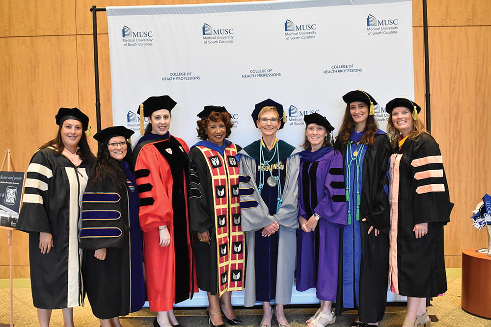 Eight people wearing faculty robes pose in front of a sign that says MUSC College of Health Professions.