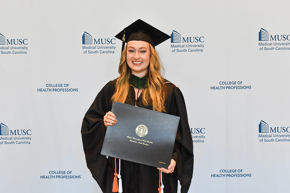 Young woman with long hair smiles while wearing a graduation robe and cap and holding a diploma.
