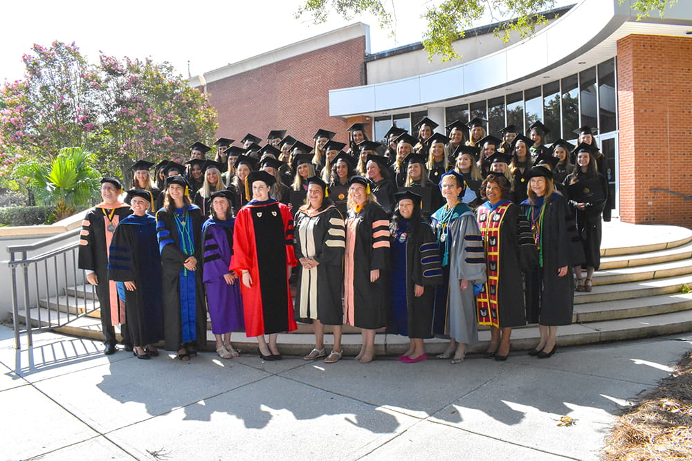 People wearing graduation robes stand in front of a brick building.