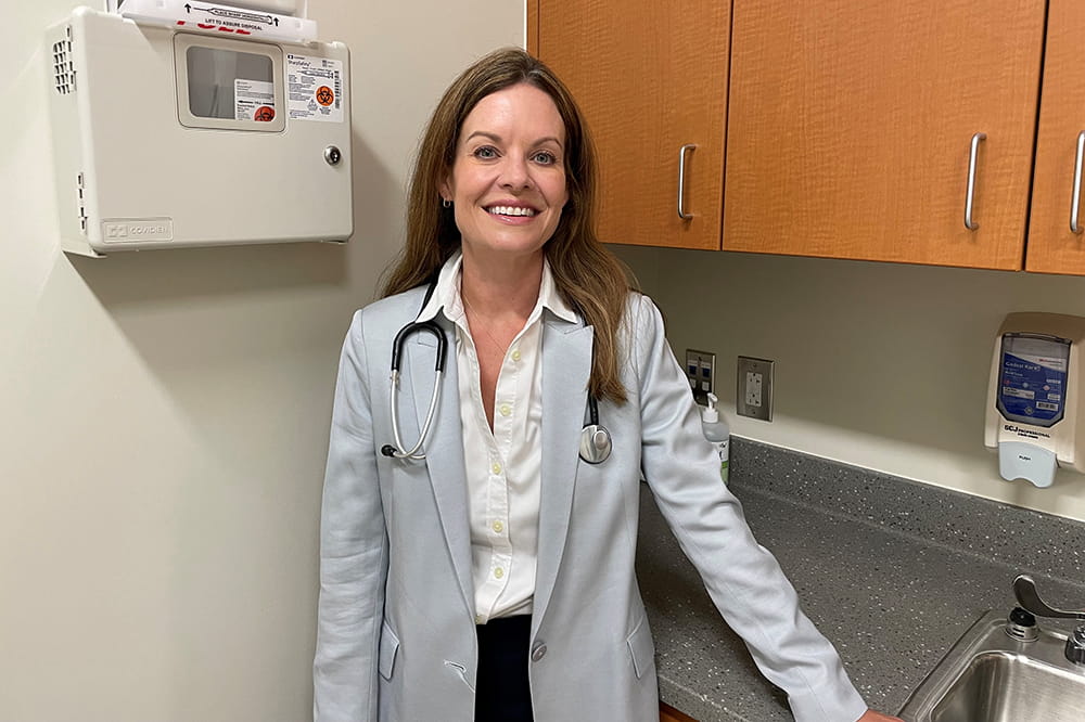 a woman in casual business attire with a stethoscope around her neck poses in an exam room