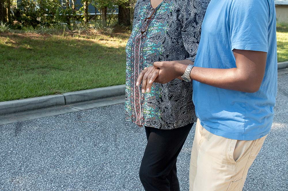 A shoulders down picture of an older woman being assisted in walking by a younger man. She is wearing a paisley blouse and black pants. He is wearing a blue t-shirt and khakis.