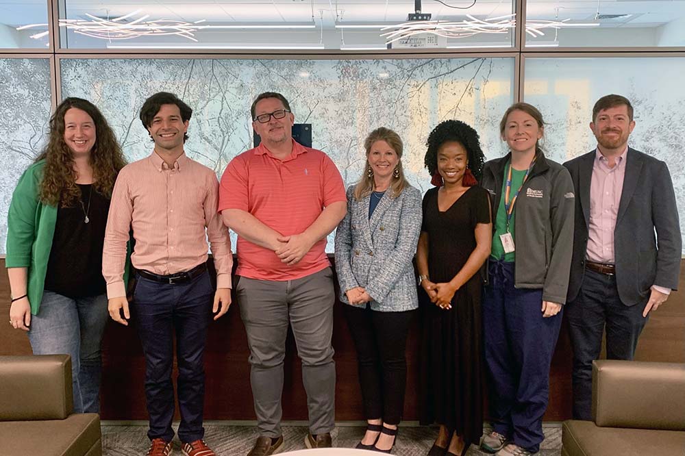 Seven people stand and smile as they post for a picture. They are dressed in business casual clothing.