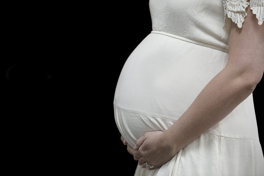 Woman cupping her pregnant belly. She is wearing a white dress with lacy sleeves.