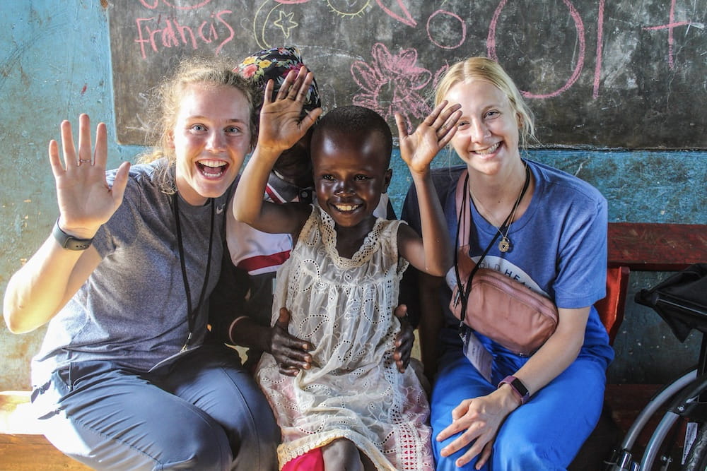 Two young female health care professionals sit on either side of a young Ugandan child who is smiling and has both hands up in the air.