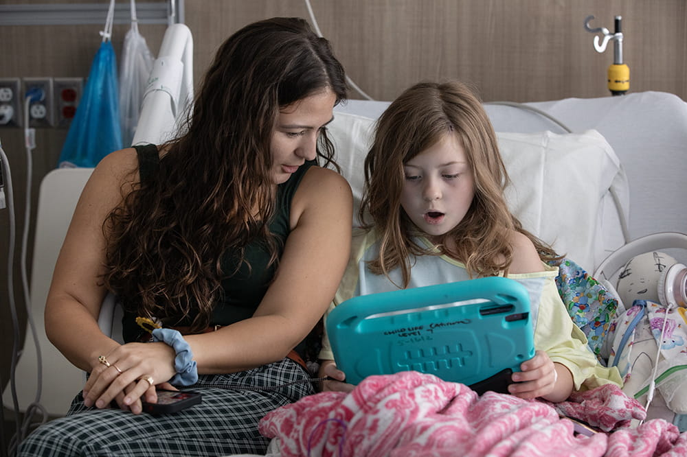 A young woman with long dark hair wearing a tank top sits on a hospital bed with a girl who has shoulder length reddish hair and is looking at a screen.
