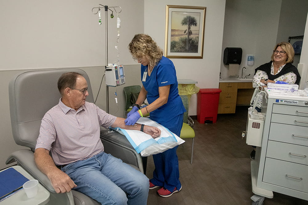  A man wearing a polo shirt and jeans sits in a chair as a woman in blue scrubs prepares to put a needle in his arm. An IV stand is beside them. A woman is standing off to the side watching.