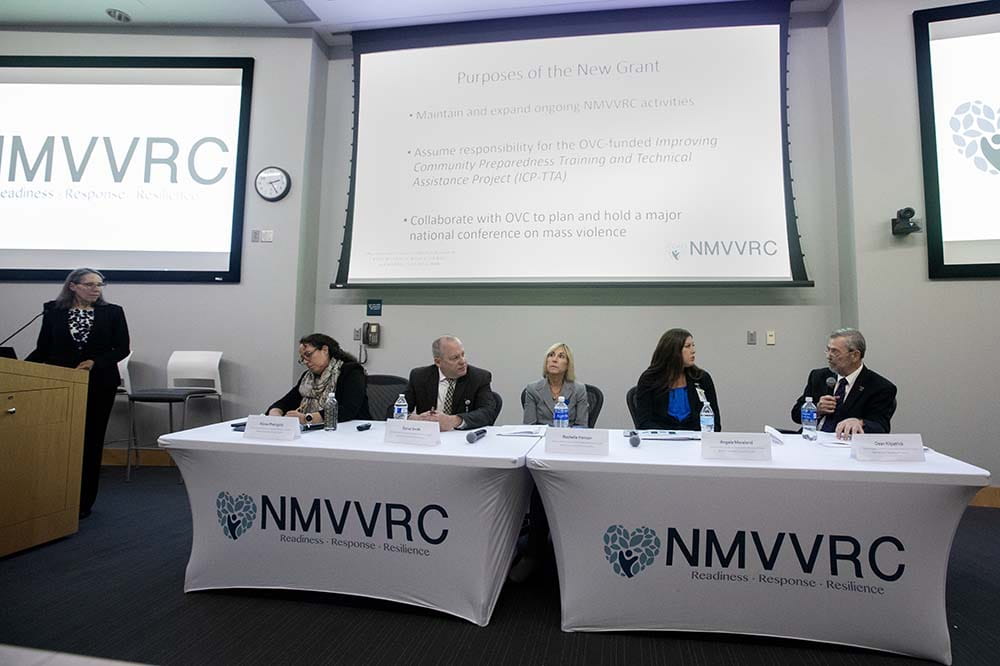 Woman stands on far left behind a podium. She's looking at two tables with the letters NMVVC on them. There are two men and three women sitting at the tables.