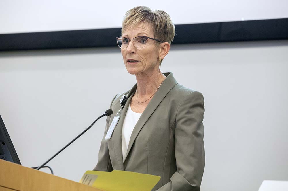 woman with short blonde hair and glasses wearing a taupe colored suit jacket speaks at a podium.