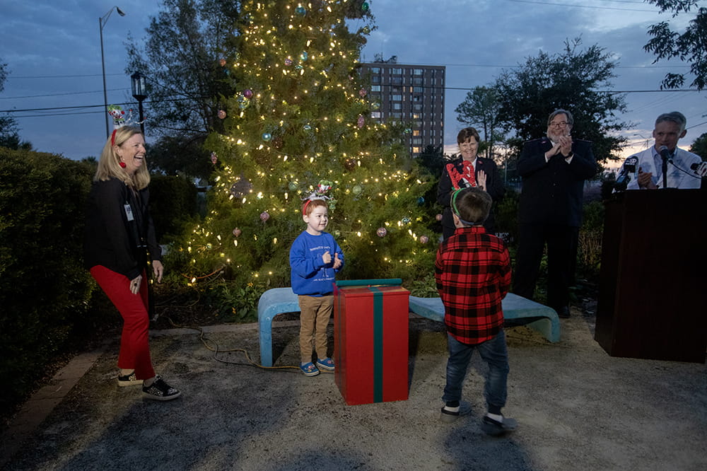 A woman laughs as two boys stand by a pretend gift. Three people and a Christmas tree are behind them.