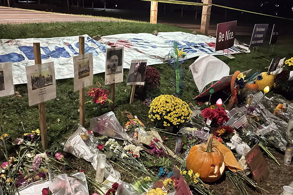 Flowers and pumpkins rest on the grass in front of signs holding photos of murder victims.