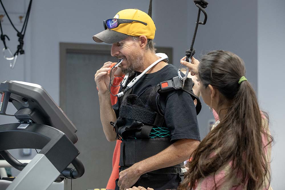 Man wearing a baseball cap puts a device in his mouth. A harness is helping to support him, as is a woman standing beside him. She has a long dark ponytail.