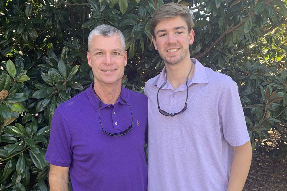 Two men wearing purple polo shirts stand together in front of greenery. The one on the left has gray hair. His son has light brown. hair. Both are smiling.