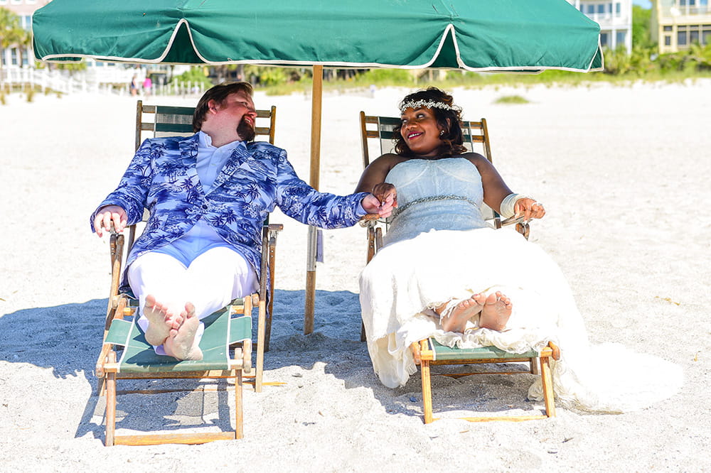 a bride and groom sit barefoot on beach chairs under a large beach umbrella