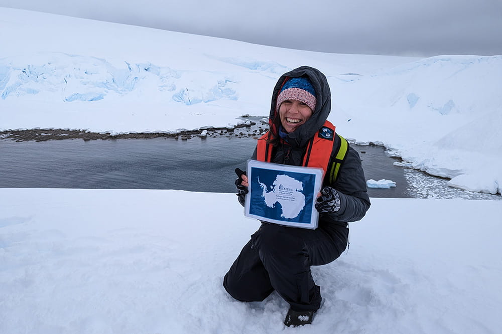 Hollings researcher Judit Jimenez Sainz kneels in Antarctica snow holding a paper with the Hollings logo
