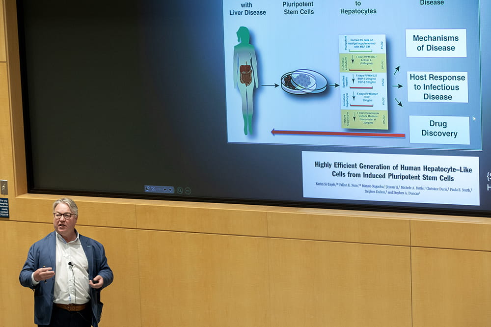 A man in a sportcoat and white shirt speaks in front of a screen that says highly efficient generation of human hepatocyte-like cells from induced pluripotent stem cells.