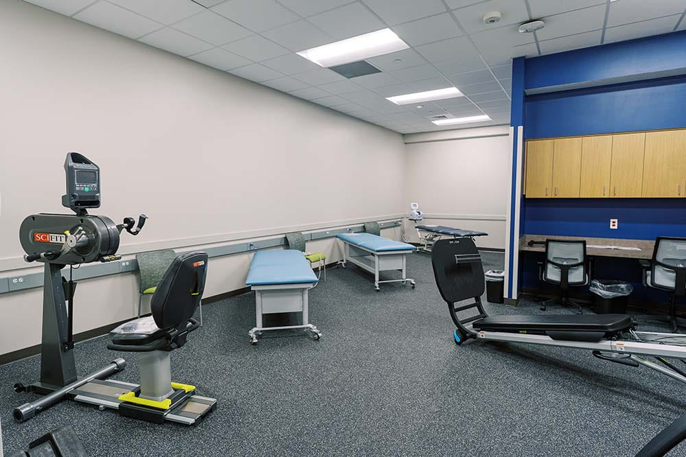 Exercise equipment and tables for therapy in a room with overhead lights.