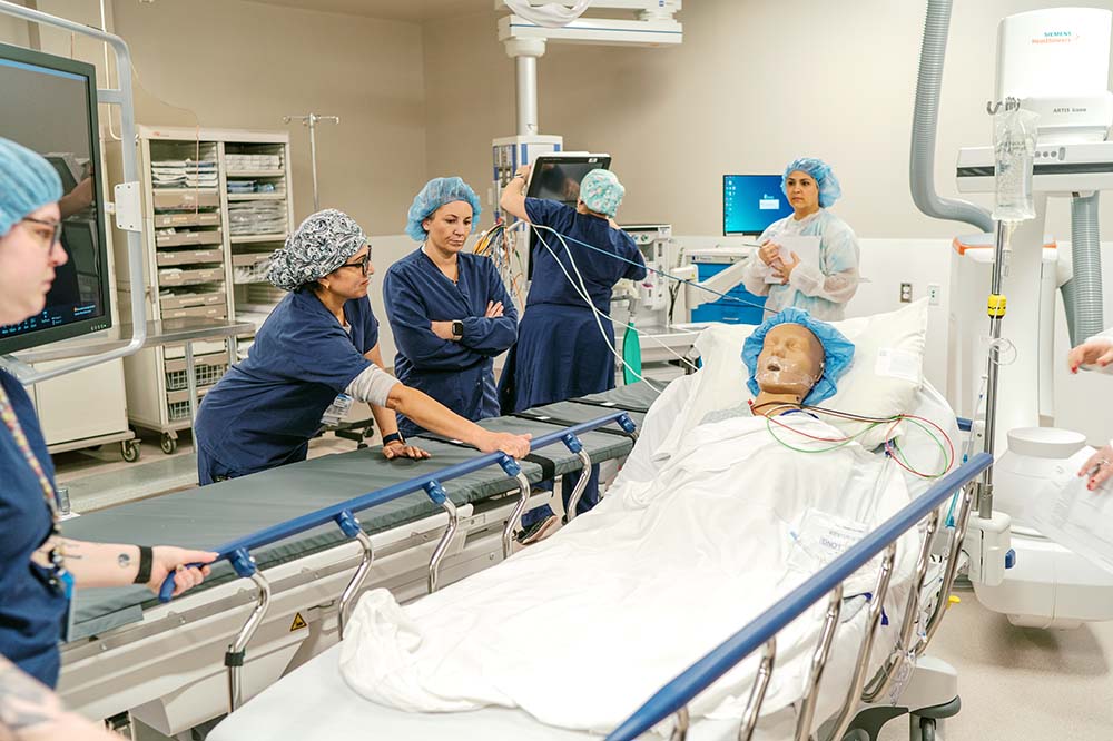 Group of women in scrubs gathered around a medical manikin who is lying on a table.