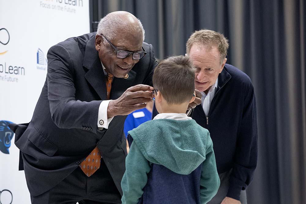 Two men in suits lean toward an elementary school student. One of the men helps the boy put on a pair of glasses.