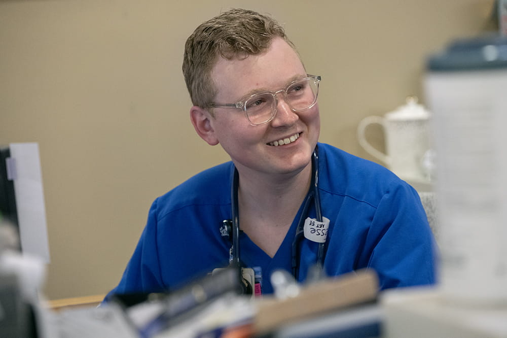 Person wearing eyeglasses and blue scrubs smiles while looking to the side.