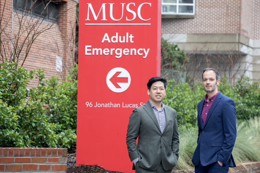 Two men in suits standing in front of a red sign that says MUSC Adult Emergency with an arrow pointing left.