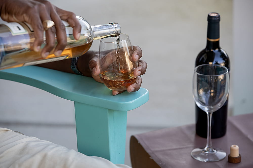 Hand wearing a ring pours brown liquid into a glass. An empty wine glass and a bottle of red wind are on a table beside the person.