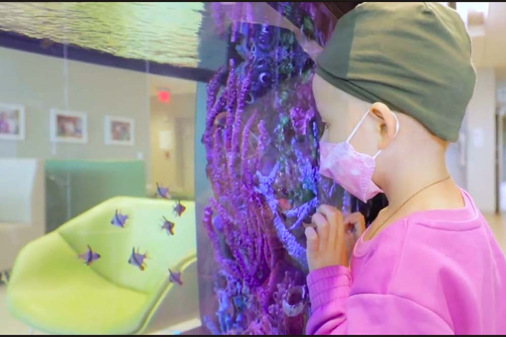 Girl wearing a headscarf, mask and pink top looks at fish in an aquarium.