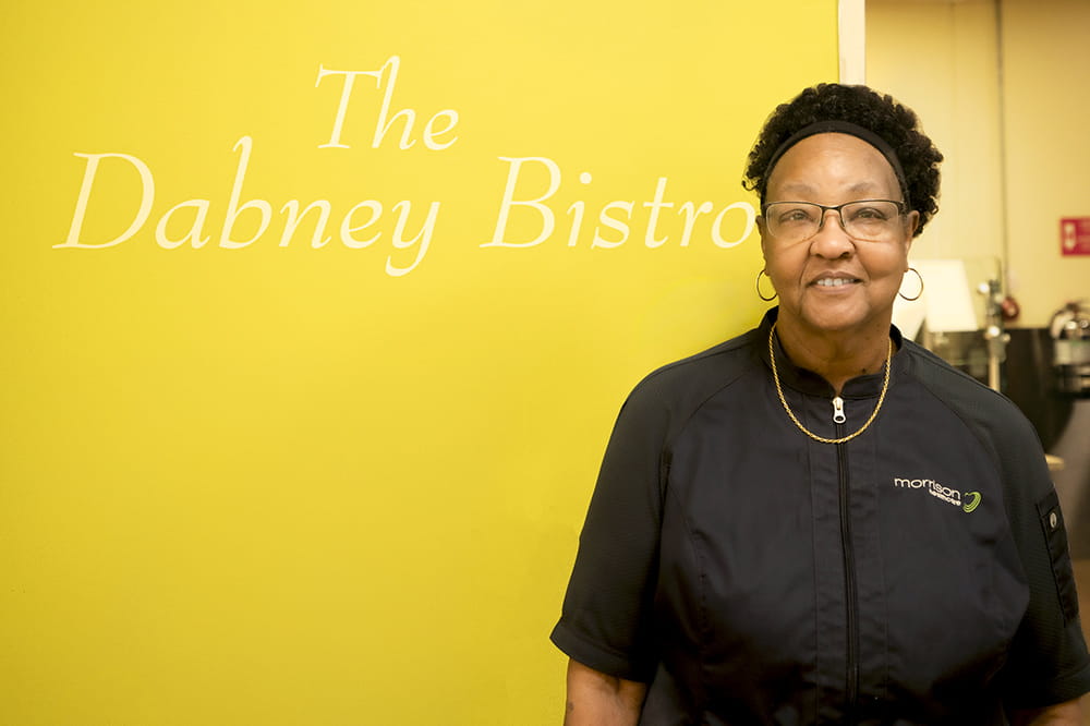 Woman wearing a work uniform smiles as she stands by a yellow wall that says The Danny Bistro.