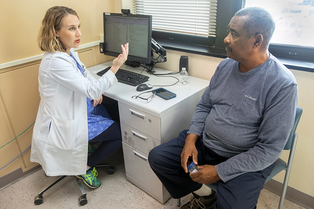 Woman in white doctor's coat gestures while talking with a seated man.
