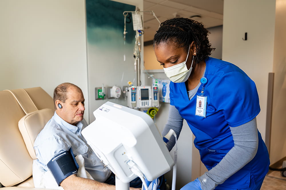 a nurse leans over a monitor while in the background a patient sits in a chair with a cuff around his arm