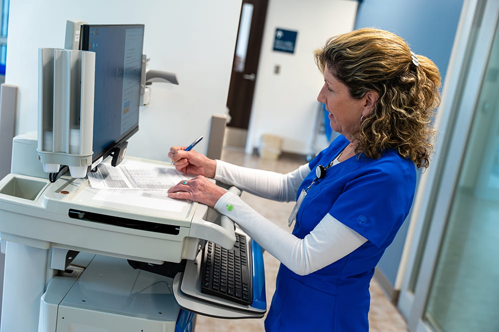 a nurse stands at a monitor in a glass-lined hallway and takes notes 