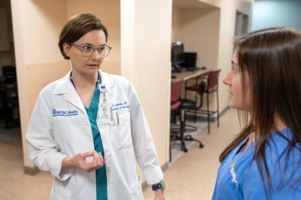 a woman in a white doctor's coat speaks with a woman in nurse's scrubs in a staff work area