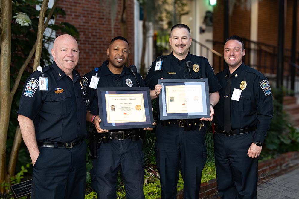 Four uniformed Public Safety officers stand in a row. Two men in the middle are holding framed awards for saving another man's life.