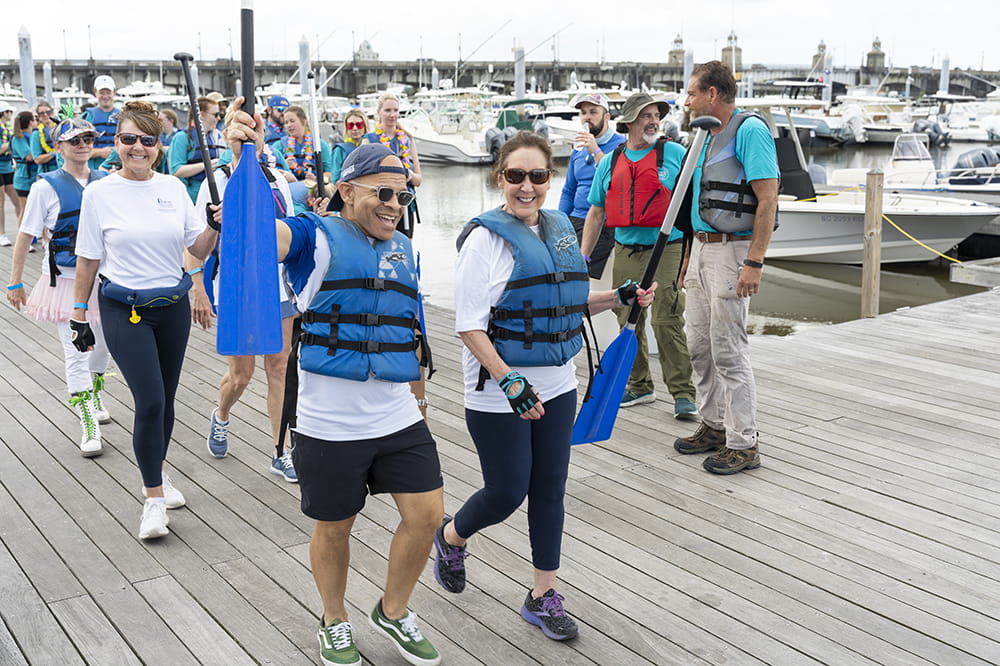 as people on a dock walk toward the water, a man in a blue life jacket with a backwards baseball cap raises his paddle in a cheer