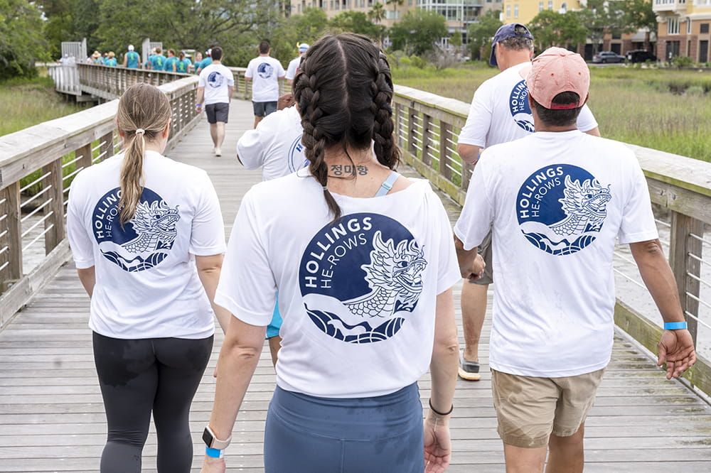 shot of the backs of people's t-shirts as they walk down a dock The white shirts with blue graphics say Hollings He-Rows with a stylized image of a dragon