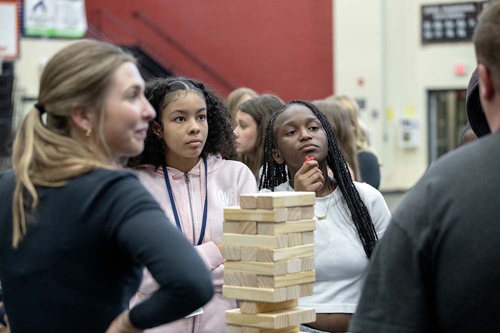 Two girls look at a person who is out of frame. A woman stands beside them. There is a Jenga tower on the table in front of them.