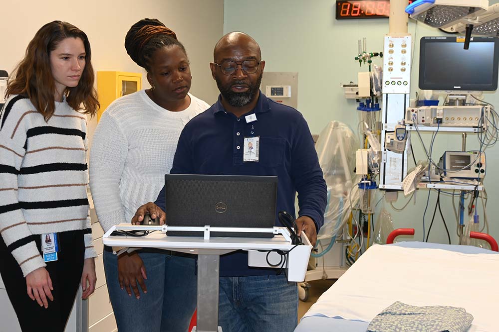 Three people stand in a hospital room. They are looking at a computer monitor.