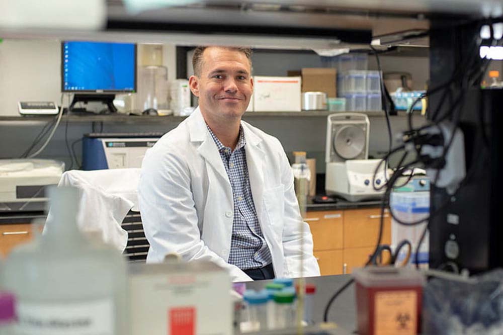 Man in a white lab coat smiles seated in a lab.