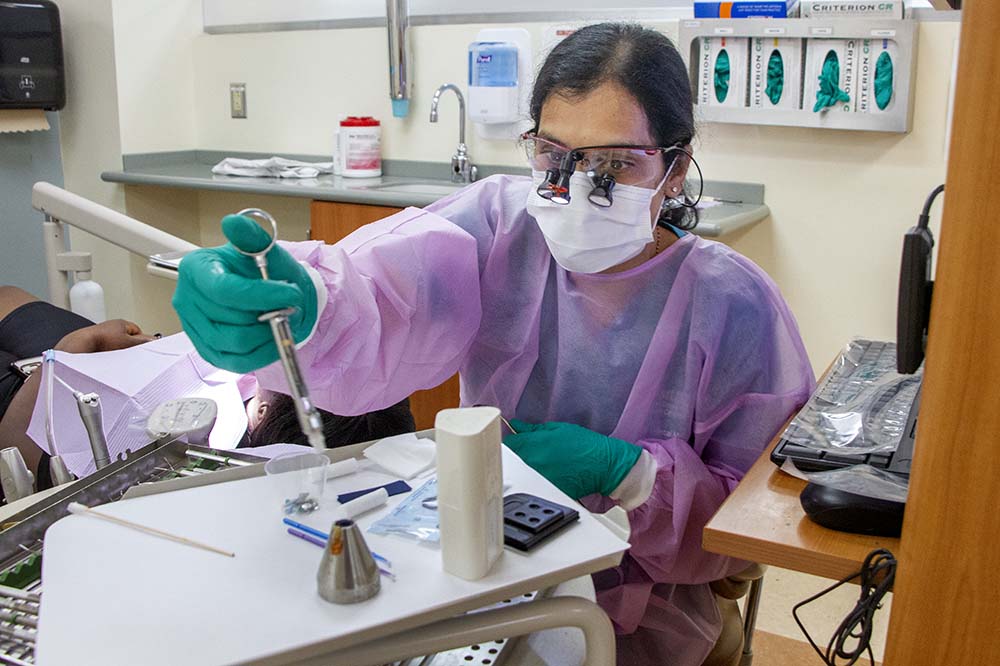 Woman wearing dental goggles and a violet colored coverup and green gloves leans forward with a dental instrument.