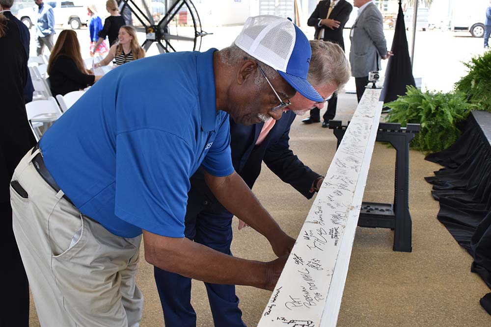 Two men sign a beam. One is wearing a baseball cap and polo style shirt. The other one is in dress clothing.
