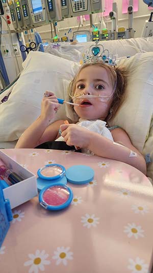 Girl in hospital bed wears tiara and has toys lying on her blanket.