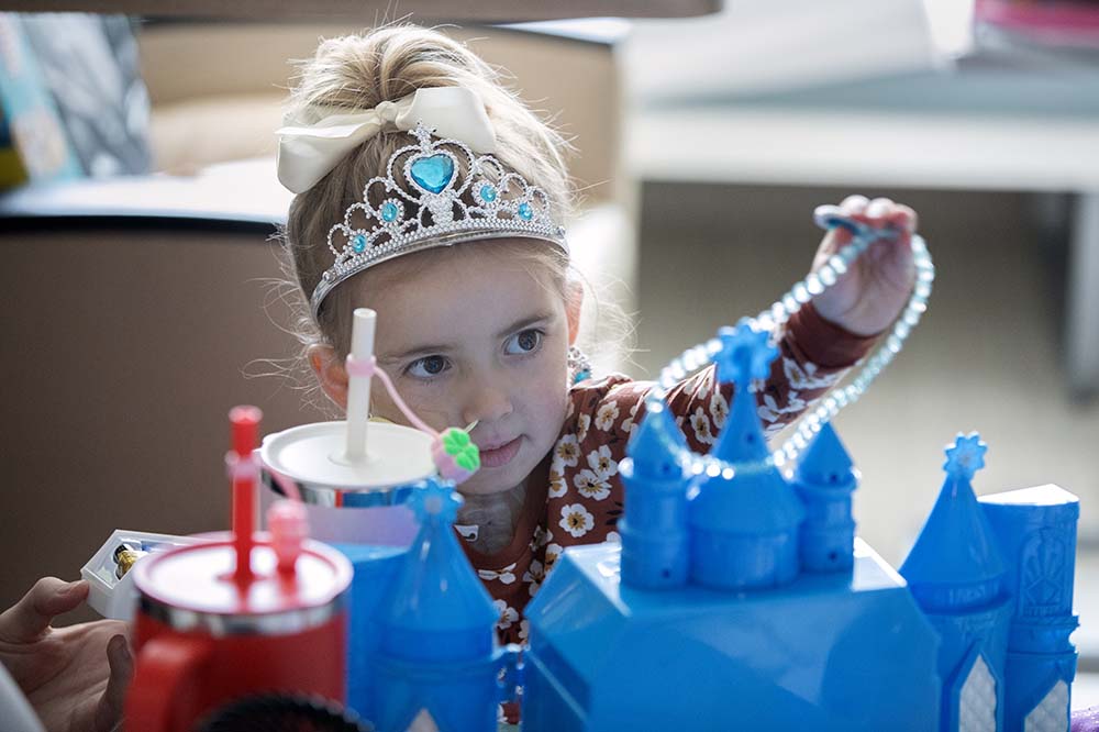 Little girl wearing a tiara plays with a necklace that is attached to a blue toy castle.