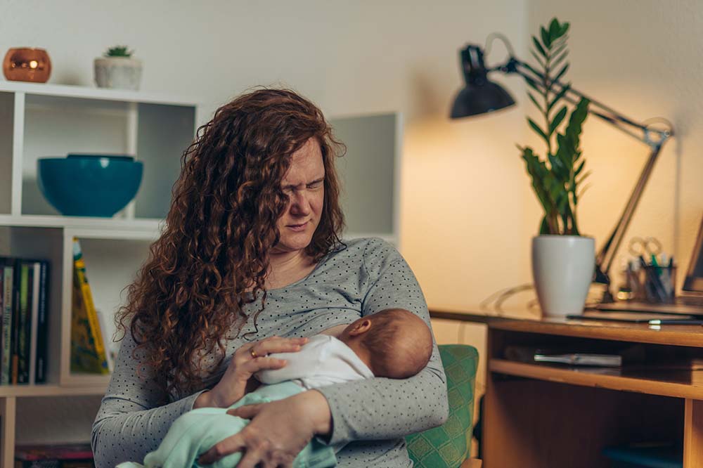 Woman with long red hair looks down at breastfeeding baby. The woman does not look happy.