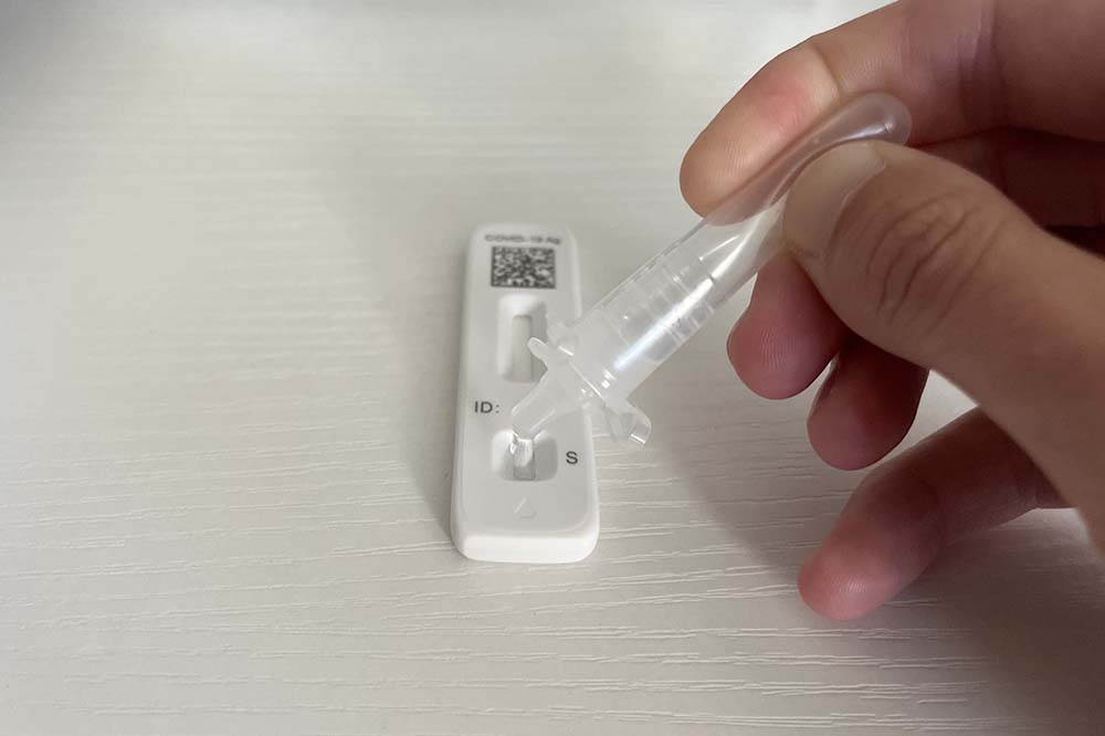 Hand holds clear colored tube to drop liquid onto a white COVID test.