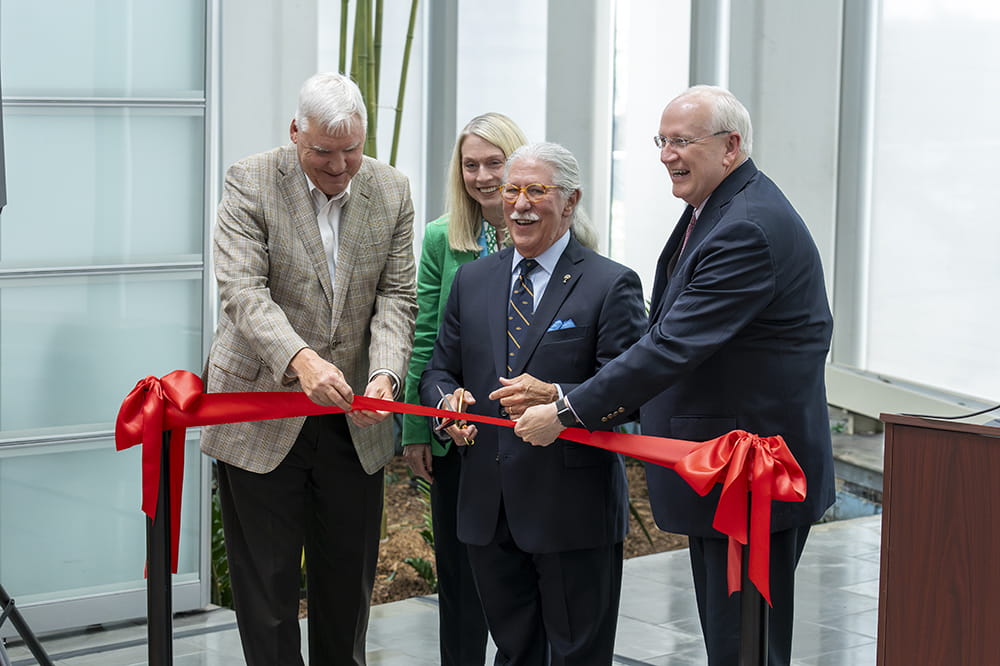 three men and a woman in suits gather around a red ribbon stretched between two poles with the main in the center wielding scissors