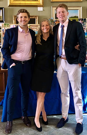 a woman with long blonde hair in a cocktail dress poses between her two adult sons, both taller than her and dressed up in blazers