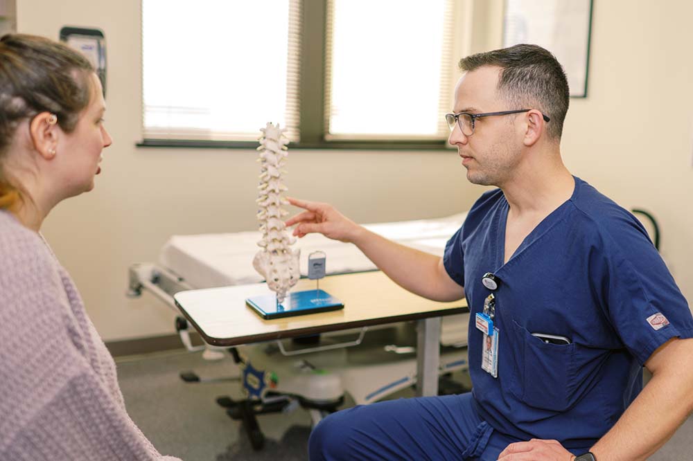 Doctor in blue scrubs points to a model of a spine as a woman watches.