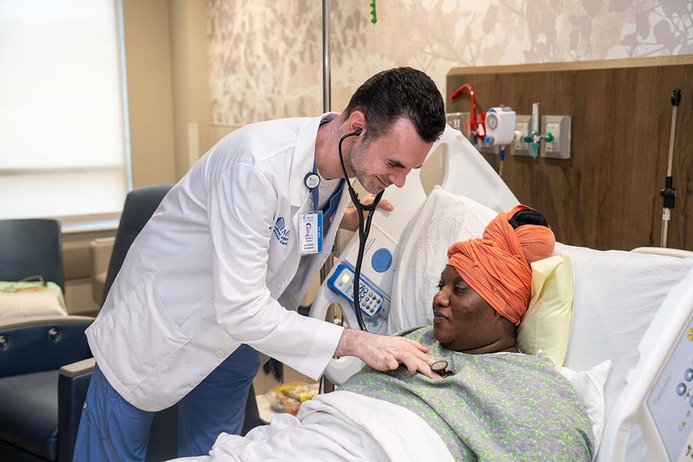 Young man leans over to use a stethoscope on a woman lying in a hospital bed.