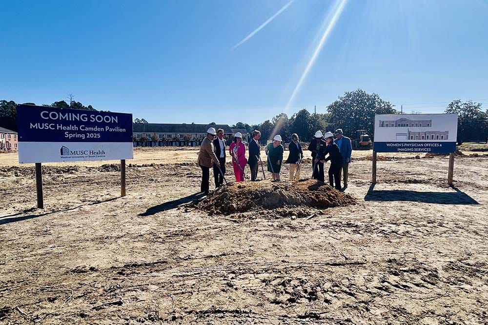 A group of people wearing hard hats stand beside two large signs on a field of dirt.